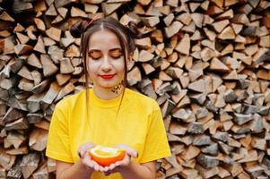 Young funny girl with bright make-up, wear on yellow shirt hold picece of orange against wooden background. photo