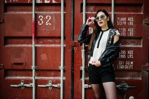 Stylish brunette girl wear on leather jacket and shorts, sunglasses against shipping container. photo