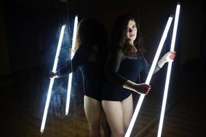 Curly brunette plus size model with two long led lamp at dance room with mirror. photo