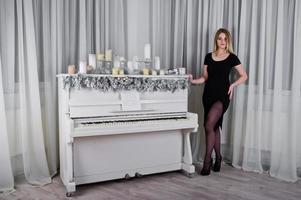 Blonde girl in black dress posed near piano with christmas candles decor in white room. photo