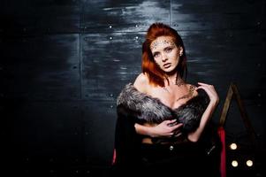 Fashion model red haired girl with originally make up like leopard predator against steel wall. Studio portrait on ladder. photo