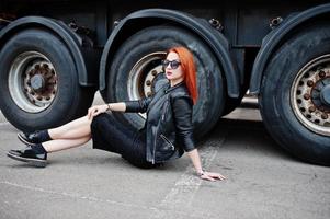 Chica elegante de pelo rojo vestida de negro, sentada contra grandes ruedas de camiones. foto