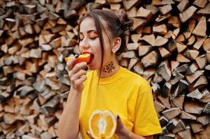 Young funny girl with bright make-up, wear on yellow shirt eat picece of orange against wooden background. photo