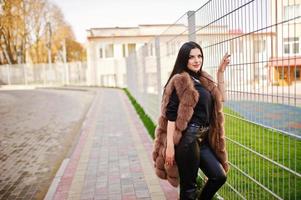 Fashion outdoor photo of gorgeous sensual woman with dark hair in elegant clothes and luxurious sleeveless fur coat against iron fence at autumn city.