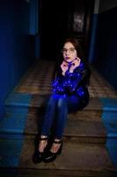 Night portrait of girl model wear on glasses, jeans and leather jacket, with blue garland on her. photo