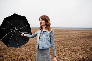 Portrait of brunette curly girl in jeans jacket with black umbrella at field. photo