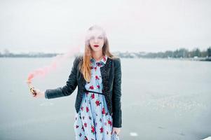 Stylish girl in leather jacket hold pink smoke flare at winter day against frozen lake. photo