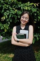 Portrait of a shy young woman in black polka dot dress and glasses holding books in the park. photo