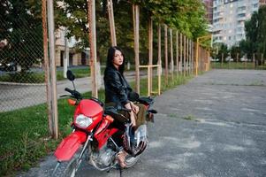 retrato de una mujer genial e impresionante con vestido y chaqueta de cuero negro sentada en una moto roja genial. foto