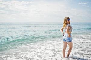 hermosa modelo relajándose en una playa de mar, usando jeans cortos, camisa de leopardo y gafas de sol. foto