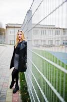 Blonde fashionable girl in long black leather coat against iron fence at street. photo