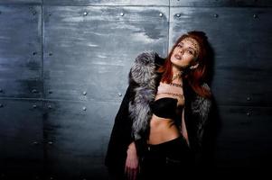 Fashion model red haired girl with originally make up like leopard predator wear on furs against steel wall. Studio portrait. photo