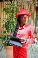 Pretty braids business african american lady bright bossy person friendly wear office red shirt, hat and trousers, hold tablet on hands. photo