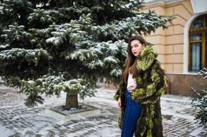 chica morena con abrigo de piel verde en el día de invierno contra un pino nevado. foto