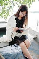 Portrait of an attractive woman wearing black polka dots dress, white shawl and glasses reading a book in a boat on a lake. photo