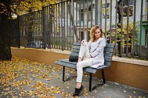 elegante modelo rubia rizada vestida de blanco con una taza de café y un teléfono móvil en las manos posando en el banco en otoño. foto