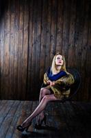 Elegante blonde girl wear on fur coat sitting on chair at studio against wooden background. photo