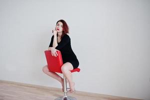 Red haired girl on black dress tunic sitting on red chair against white wall at empty room. photo