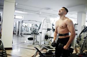Muscular arab man training in with dumbbells modern gym. Fitness arabian men with naked torso doing workout . photo