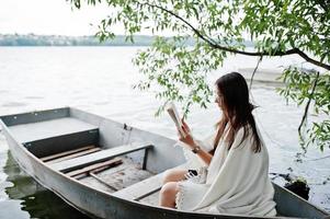 Portrait of an attractive woman wearing black polka dots dress, white shawl and glasses reading a book in a boat on a lake. photo