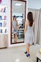 Portrait of a fantastic young woman in grey dress trying on new shoes and handbag in front of a mirror in shop. photo