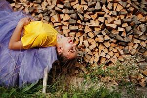 Young funny girl with bright make-up, like fairytale princess, wear on yellow shirt and violet skirt lying against wooden background. photo