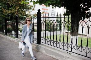 Girl in gray coat with sunglasses and handbag walking at street against iron fence. photo