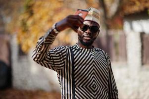 Portrait of stylish black african american man at hat and sunglasses against sunny autumn fall background doing selfie from mobile phone. Rich people in africa at traditional dress. photo