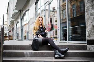 chica rubia de moda con un largo abrigo de cuero negro posado en las escaleras. foto