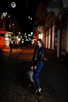 Night portrait of girl model wear on glasses, jeans and leather jacket, with backpack , against lights of city streets. photo