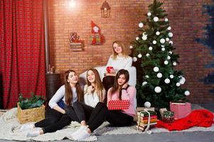 cuatro lindas amigas usan suéteres cálidos, pantalones negros y sombreros de santa contra el árbol de año nuevo con decoración navideña en el estudio. foto