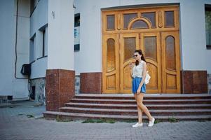 Curly stylish girl wear on blue jeans skirt, blouse and listening music from headphones phone. Portrait on streets of city. photo