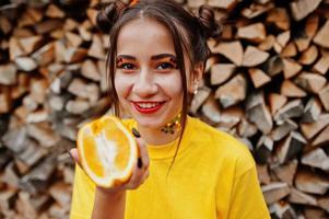 Young funny girl with bright make-up, wear on yellow shirt hold picece of orange against wooden background. photo