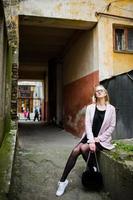 Blonde girl at glasses and pink coat, black tunic and handbag posed at street. photo