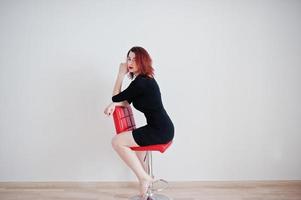 Red haired girl on black dress tunic sitting on red chair against white wall at empty room. photo