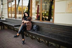 Fashion outdoor photo of gorgeous sensual woman with dark hair in elegant clothes and luxurious sleeveless fur coat and with backpack sitting on bench at autumn city.