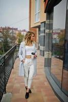 elegante modelo rubia rizada vestida de blanco con una taza de café en la mano posando contra una ventana grande. foto