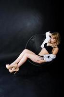 Studio portrait of sexy blonde girl with originally make up on neck and tattoo on thigh, wear on black underwear at dark background, sitting on chair. photo