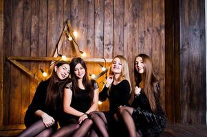 Four cute friends girls wear black dresses against large light christmas star decoration on wooden background. photo