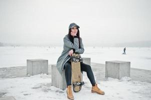 chica morena elegante con gorra gris, estilo casual de calle con patineta en el día de invierno. foto