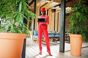 Pretty braids business african american lady bright bossy person friendly wear office red shirt, hat and trousers, hold tablet on hands. photo