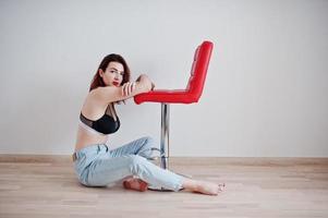 Red haired girl with a big bust on black bra and jeans sitting near red chair against white wall at empty room. photo