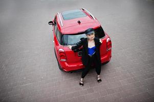 Portrait of beautiful blonde sexy fashion woman model in cap and in all black, blue corset, with bright makeup near red city car. photo