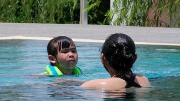 menina em um colete salva-vidas tem medo de nadar na piscina profunda. mãe ensinando filha a praticar natação na piscina. família feliz, mãe e filha brincando na piscina. video