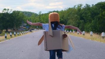 Cute dreamer little girl running on a lakeside road with a cardboard plane on a sunny day. Happy kid playing with cardboard plane against blue sky background. Childhood dream imagination concept. video