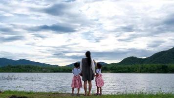 feliz madre camina de la mano con lindas hermanitas disfrutando de la naturaleza junto al lago. familia feliz, madre y sus hijas jugando sintiéndose felices en el parque. video