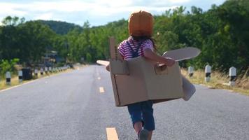 Cute dreamer little girl running on a lakeside road with a cardboard plane on a sunny day. Happy kid playing with cardboard plane against blue sky background. Childhood dream imagination concept. video