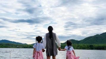 la madre felice cammina mano nella mano con le sorelline carine che si godono la natura in riva al lago. famiglia felice, madre e le sue figlie che giocano sentendosi felici nel parco. video