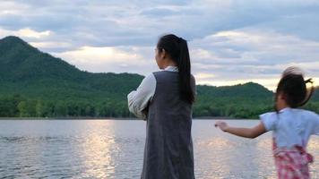 la bambina sveglia correva abbracciando sua madre da dietro mentre si godeva la vista della natura in riva al lago al tramonto. famiglia felice, madre e figlia che giocano sentendosi felici nel parco. video