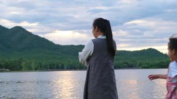 la bambina sveglia correva abbracciando sua madre da dietro mentre si godeva la vista della natura in riva al lago al tramonto. famiglia felice, madre e figlia che giocano sentendosi felici nel parco. video